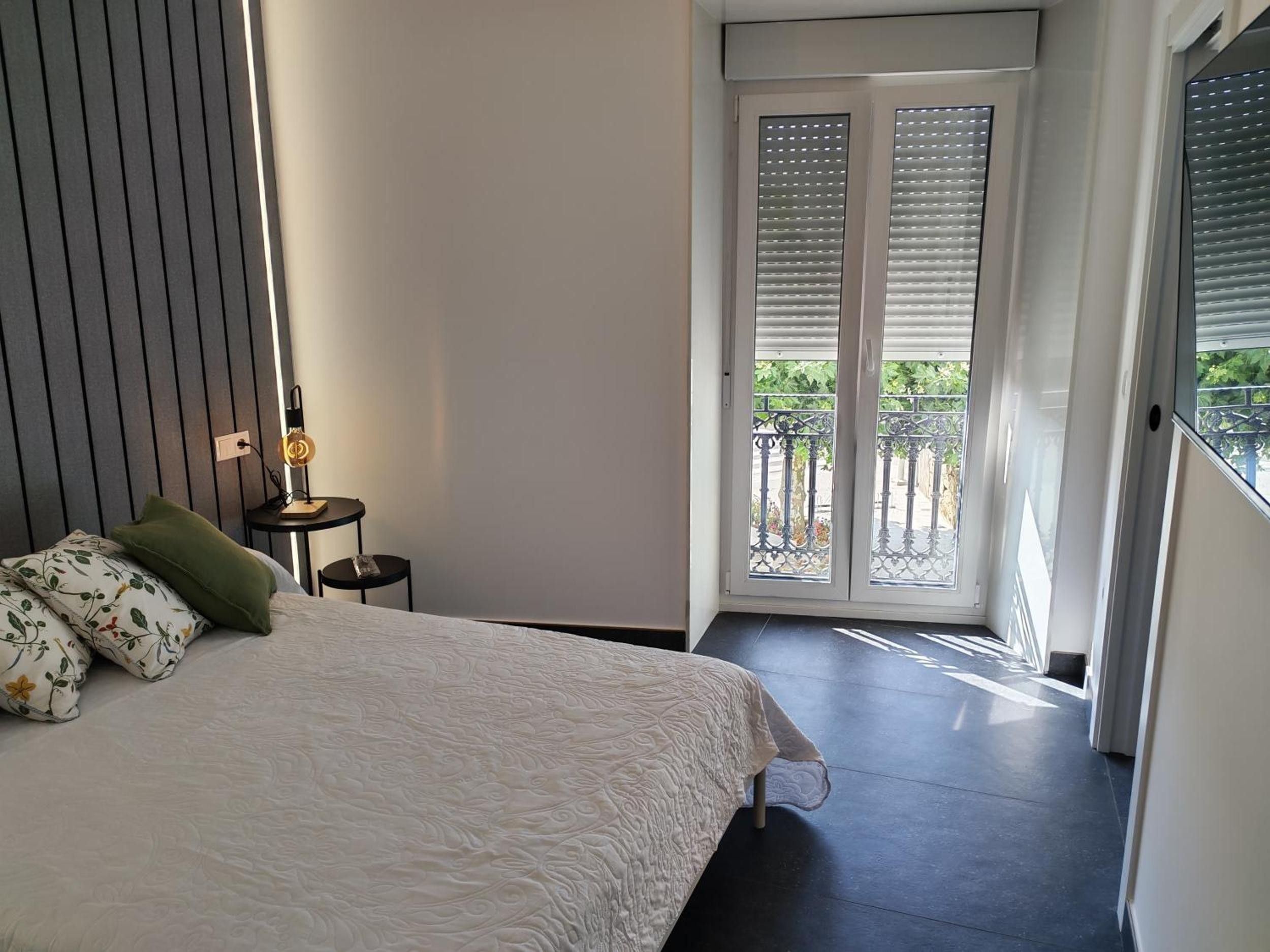 Un dormitorio con cama, mesita de noche con lámpara, ventana con persianas y vista al exterior.