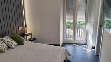 Un dormitorio con cama, mesita de noche con lámpara, ventana con persianas y vista al exterior.