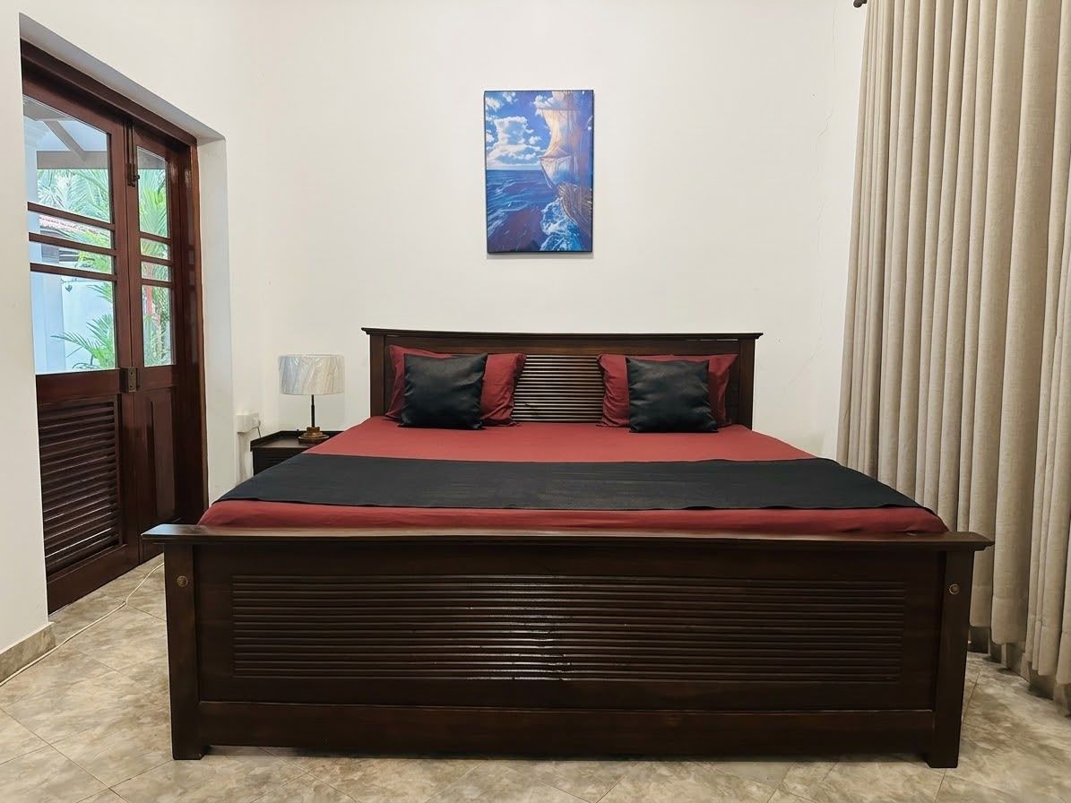A large bed with a dark headboard, a red bedspread, and two pillows. There is a painting on the wall above the bed.