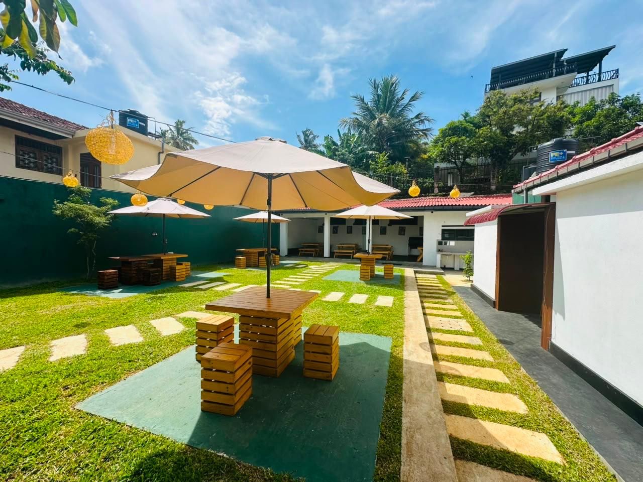 An outdoor patio area with tables and chairs, surrounded by a green lawn and a pathway.