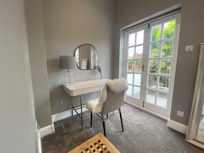 Ein Zimmer mit einem weißen Schminktisch, einem runden Spiegel, einer Lampe, einem flauschigen Sessel und Blick auf Grün durch ein großes Fenster.