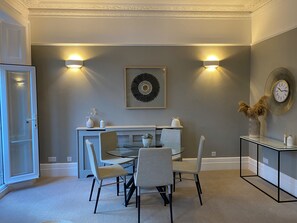 A dining area with a round table, four chairs, a wall-mounted clock, and two wall sconces. - Lovely large one bedroom flat with a balcony (London)