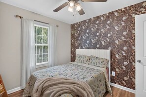 Ein Schlafzimmer mit einem Bett, einem Deckenventilator, einem Fenster mit Vorhängen und einer Wand mit Blumenmuster.