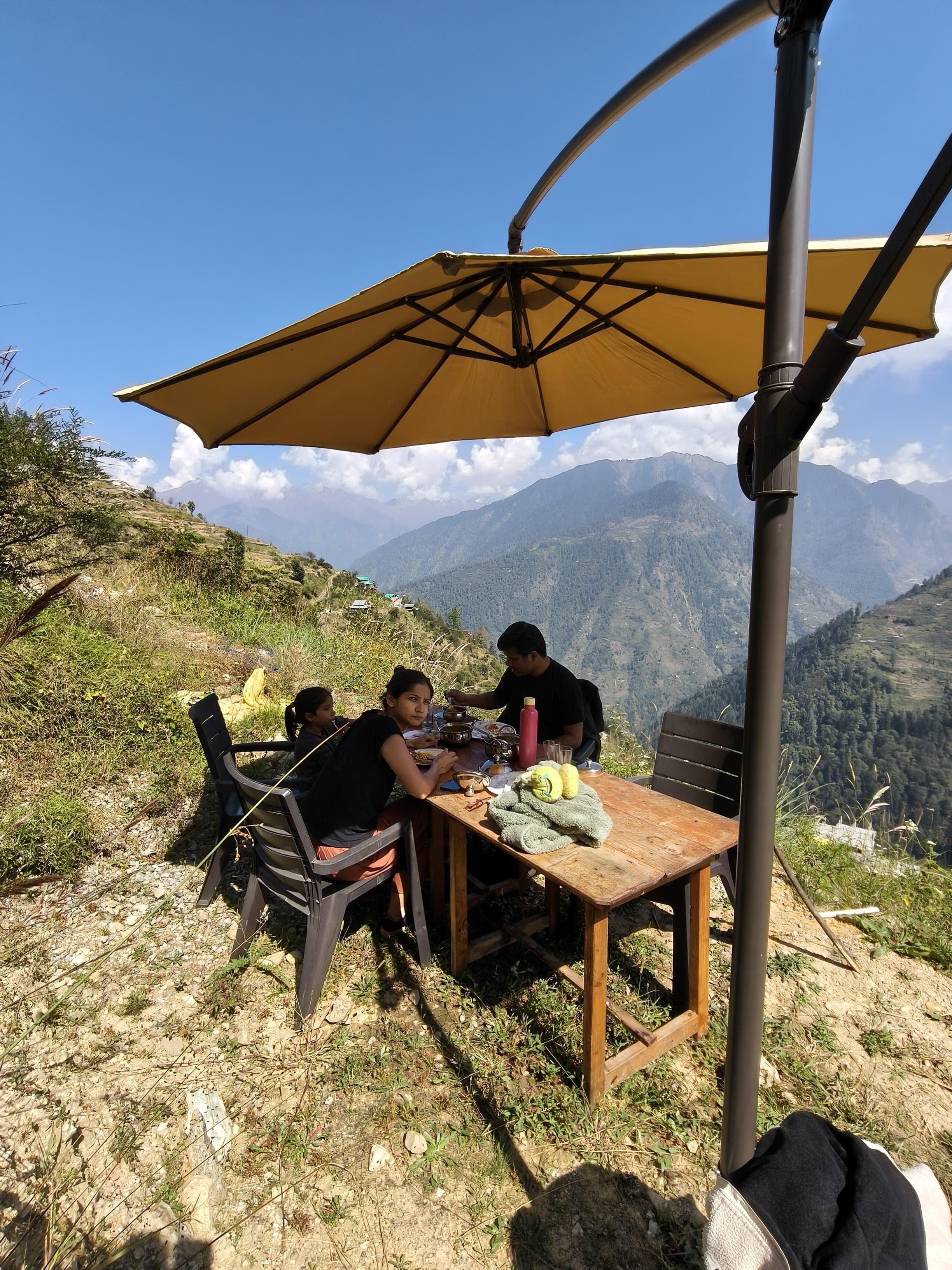 A group of people dining at an outdoor table with a large umbrella, surrounded by a mountainous landscape.