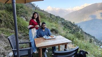 Une table avec une assiette de nourriture, deux chaises, un parasol et un chien, le tout installé sur une colline avec vue sur la montagne.