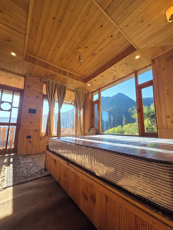 A wooden cabin room with a large bed, wooden walls, and a view of mountains through the windows.