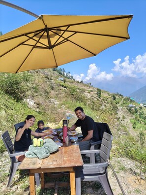 A wooden table with a yellow umbrella, plastic chairs, and a meal setting outdoors with a mountainous backdrop. - The Tirthan Valley Wooden House  (Banjar)