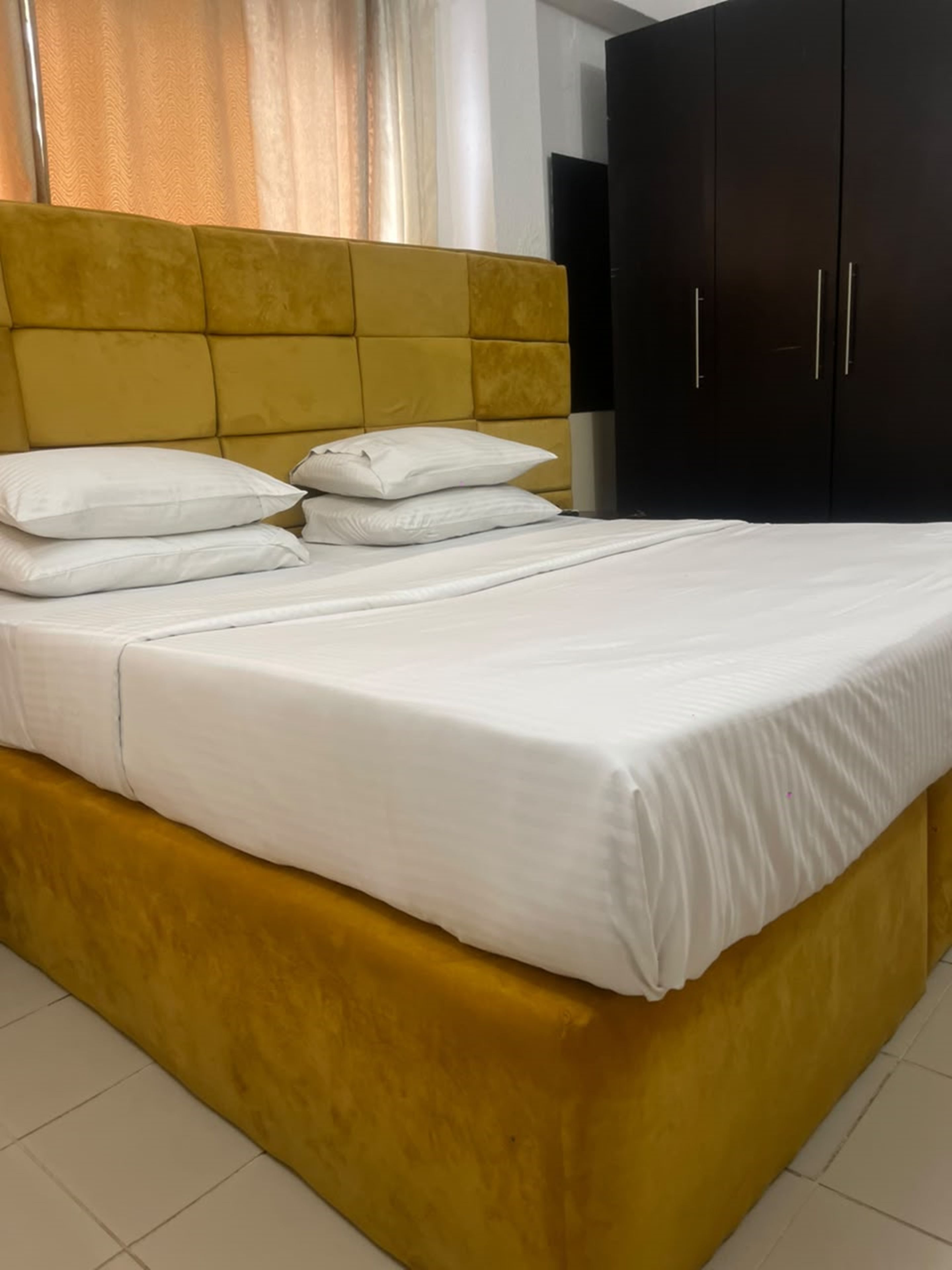 A bed with white bedding and pillows, a yellow headboard, and a dark wardrobe in a room with white walls and a tiled floor.