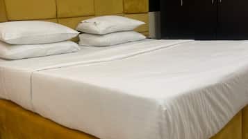 Una cama con ropa blanca y almohadas, un cabecero amarillo y un armario oscuro en una habitación con paredes blancas y suelo de baldosas.