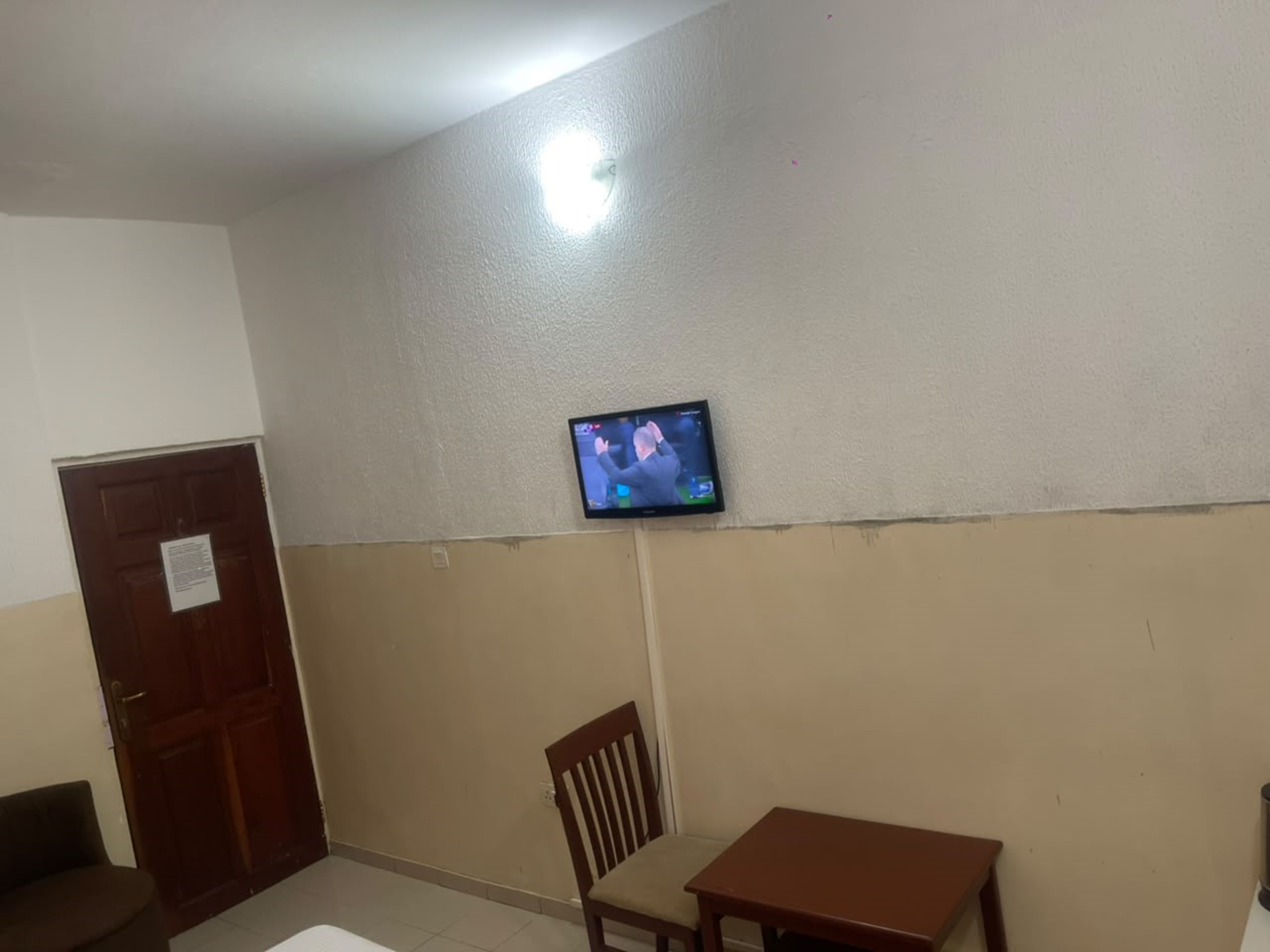 A room with a television mounted on the wall, a wooden door, a chair, and a small table.
