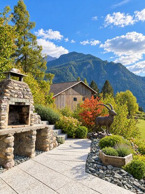 A rustic stone structure with a fireplace, a wooden building with a sloped roof, and a mountain backdrop. - Brienz Mountain Base – Simple & Comfortable (Brienz/Brinzauls)