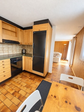 A compact kitchen with wooden cabinets, a black refrigerator, a stove, and a dining area with a table and chairs. - Brienz Mountain Base – Simple & Comfortable (Brienz/Brinzauls)