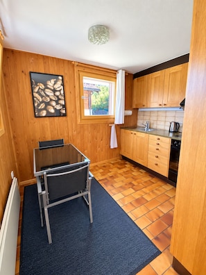 A compact kitchen with wooden cabinets, a window with curtains, a small table with chairs, and a tiled floor. - Brienz Mountain Base – Simple & Comfortable (Brienz/Brinzauls)