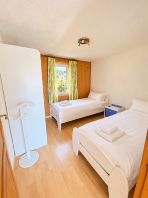A room with two beds, a window with curtains, a bedside table, and a lamp. - Brienz Mountain Base – Simple & Comfortable (Brienz/Brinzauls)