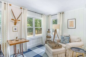 Ein Zimmer mit Schachbrettmuster-Boden, einem Holztisch mit Schachfiguren, einem Sofa mit blauem Kissen, einer Lampe und einem an der Wand befestigten Skulpturenkopf eines Giraffen.