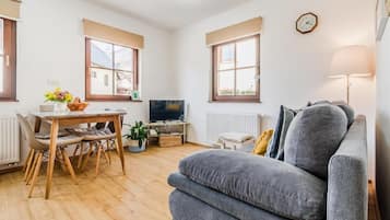 Una sala de estar acogedora con un sofá gris, una mesa de comedor de madera con sillas, un televisor y un ventanal con vistas.