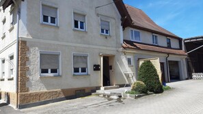Un edificio de dos pisos con fachada de piedra, múltiples ventanas con contraventanas y un pequeño jardín delantero con un arbusto cuidado.