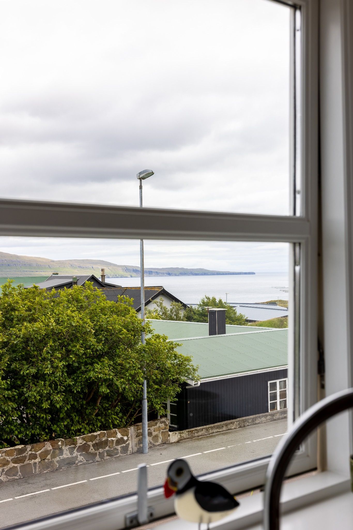 A view through a window of a building with a stone wall, a road, and a body of water in the distance.