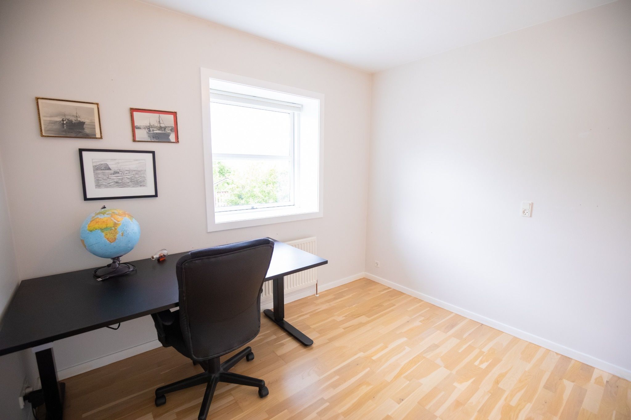 A room with a wooden floor, a black desk with a globe, a chair, and framed pictures on the wall.