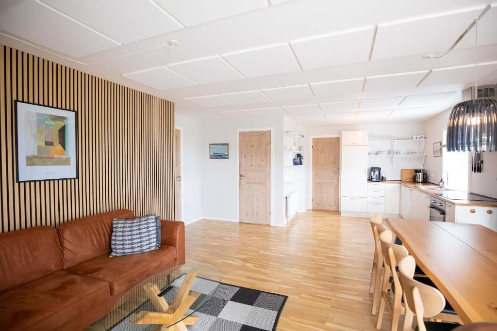 Un espace de vie moderne avec un mur à lattes de bois, un canapé en cuir marron, une table basse en verre, une table à manger en bois avec des chaises, et un coin cuisine avec des meubles et des appareils électroménagers blancs.