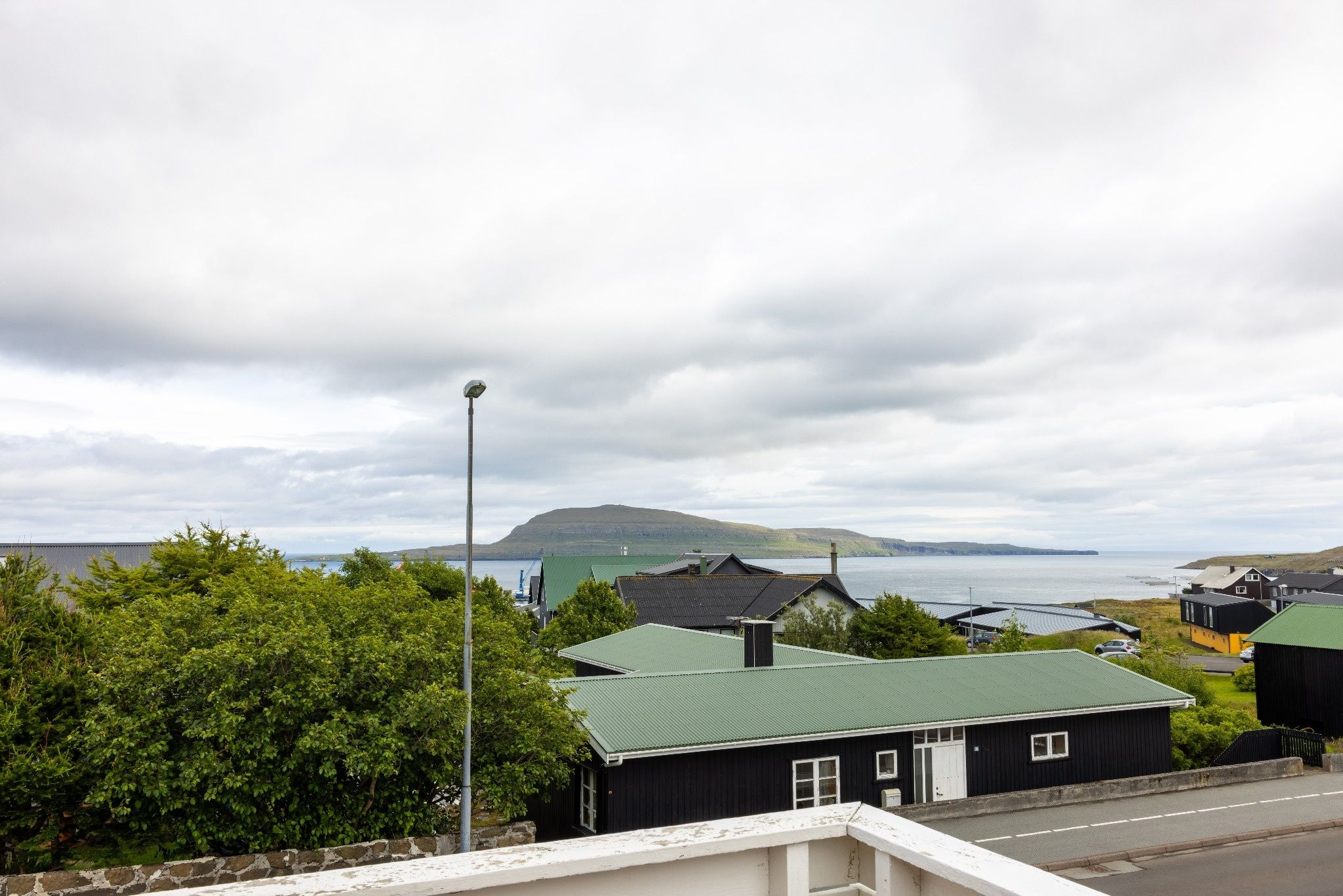 A coastal village with houses, a street lamp, and a view of the sea and hills.