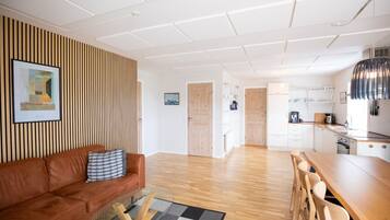 A modern living area with a sofa, coffee table, and a dining table with chairs. There is a kitchen visible in the background.