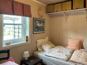 Une chambre à coucher avec un lit, une table de chevet, une lampe, une fenêtre donnant sur un beau paysage, un tableau et une étagère avec des paniers.