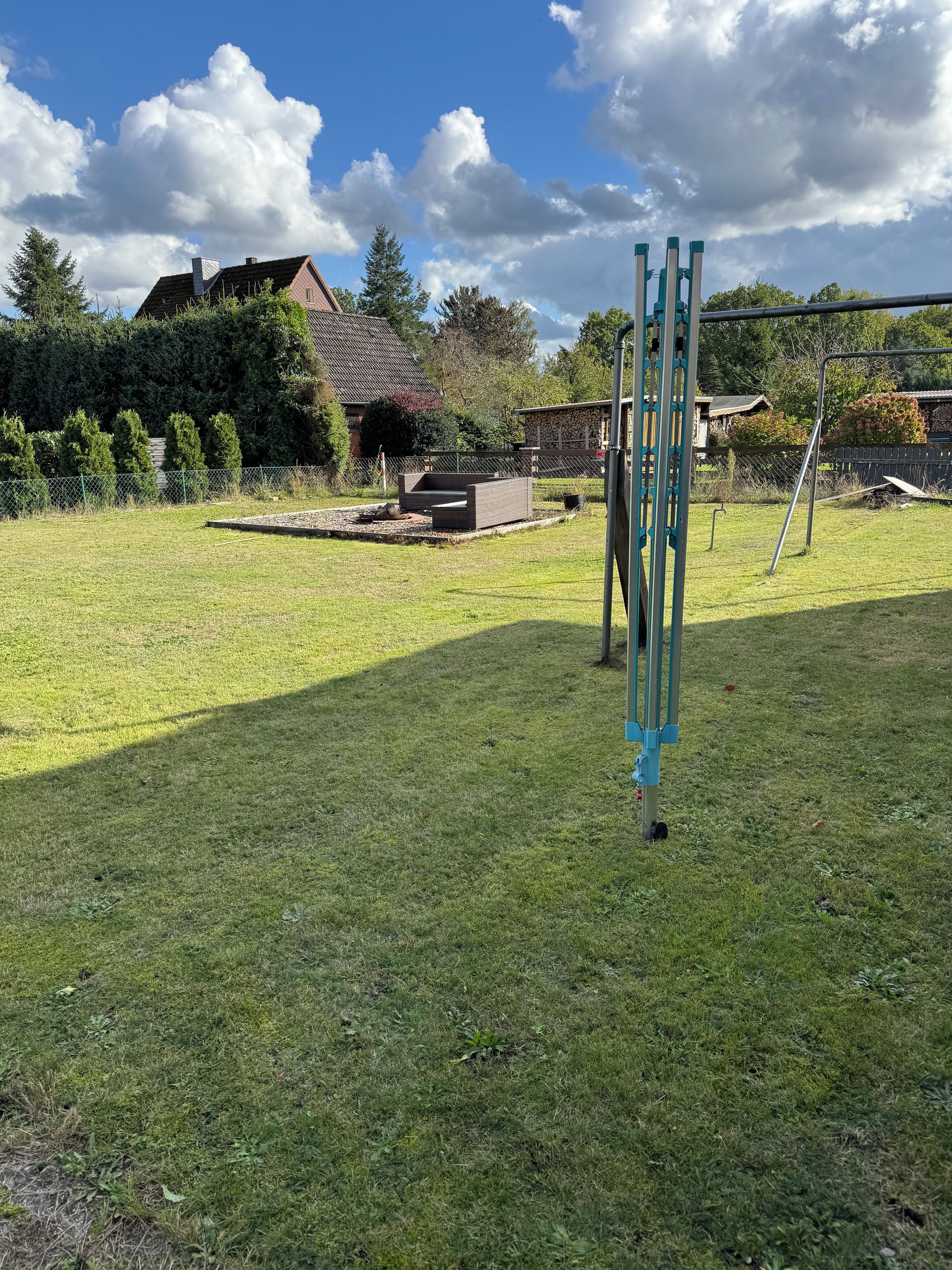 Ein grasbewachsenes Freigelände mit einer Metallkonstruktion, einer Steinmauer und einem Haus im Hintergrund.