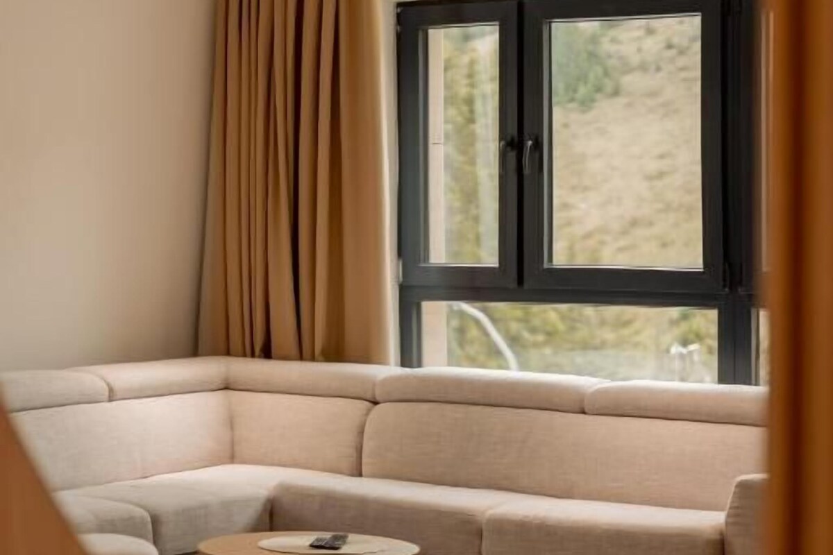 Ein modernes Wohnzimmer mit einem beigen Ecksofa, einem kleinen runden Couchtisch und einem großen Fenster mit Vorhängen.