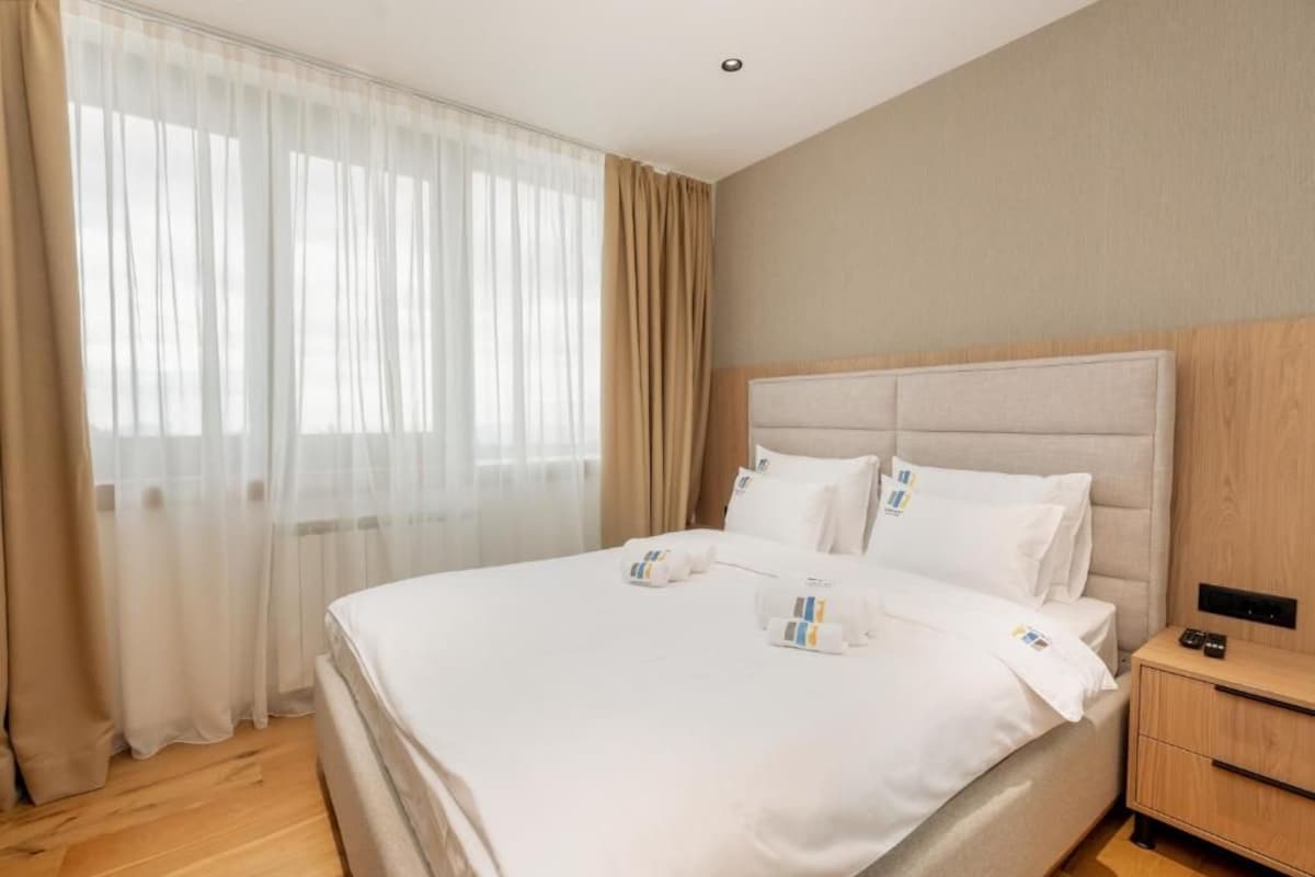 Ein Hotelzimmer mit einem großen Bett, einem Nachttisch aus Holz und einem Fenster mit durchscheinenden Vorhängen.
