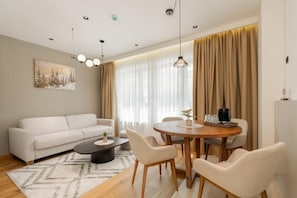 Ein modernes Wohnzimmer mit einer Couch, einem Couchtisch, einem Esstisch und Stühlen. An der Wand hängt ein Gemälde, und ein Fenster ist mit Vorhängen versehen.