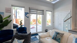 A living room with a white sofa, a blue armchair, and a coffee table. There are two windows with blinds, a door, and a staircase. - New 3br Townhome, Heated Pool, Beach Steps Spi 3 Bedroom Townhouse (South Padre Island)