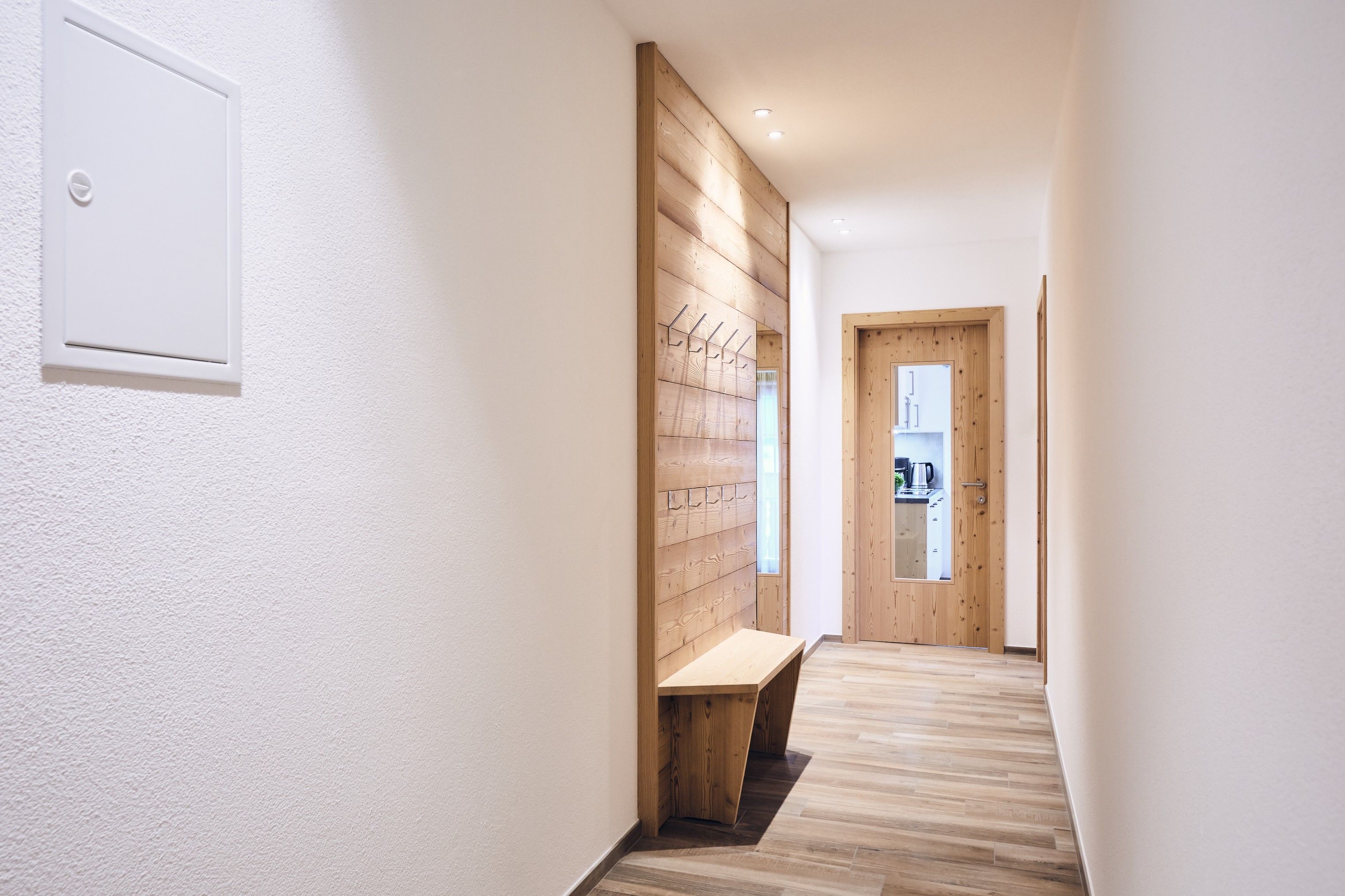 A hallway with a wooden bench, a coat rack, and a door leading to a room with a kitchenette.