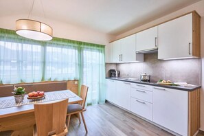 A modern kitchen with white cabinets, a wooden dining table, and a window with green curtains. - Apartment 'Huberhof - Puschtra' (Santo Stefano)