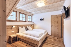 A bedroom with a wooden bed, a nightstand, a wall-mounted TV, and a window with a view of a mountain. - Apartment 'Huberhof - Puschtra' (Santo Stefano)