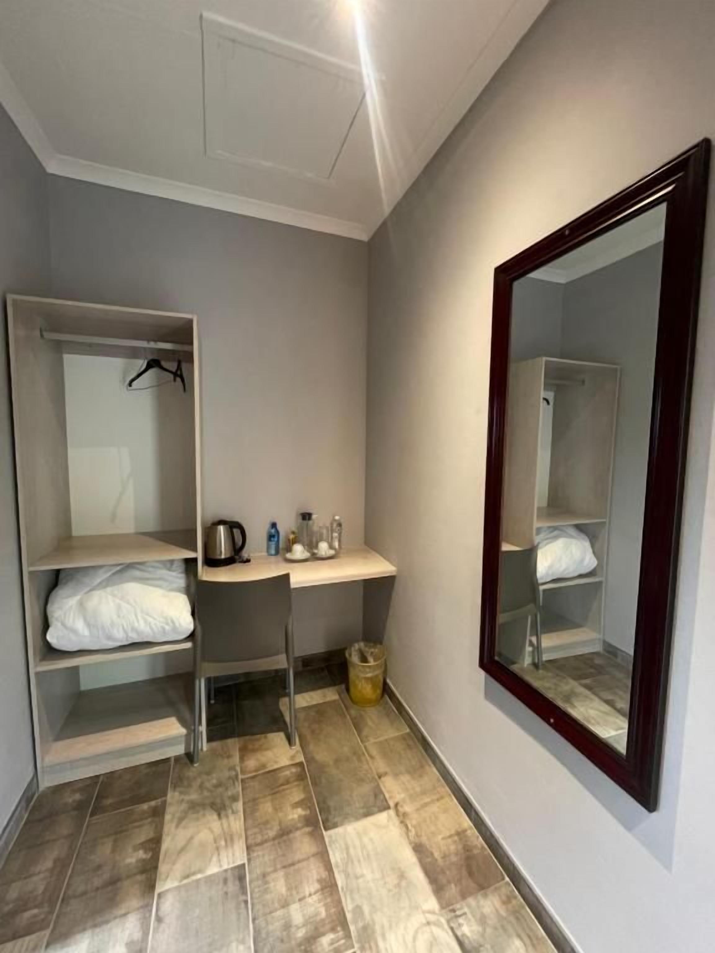 Una habitación pequeña con una cama, un escritorio, una silla, un espejo y una estantería con una tetera y botellas.