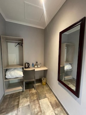 Una habitación pequeña con una cama, un escritorio, una silla, un espejo y una estantería con una tetera y botellas.