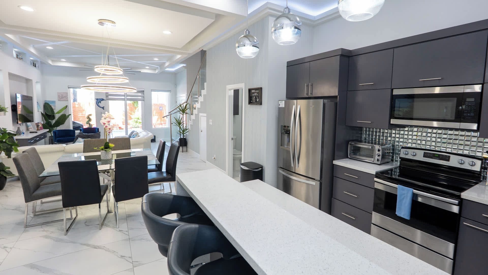 A modern kitchen with stainless steel appliances, a dining area with a glass table, and a living space with a sofa and plants.