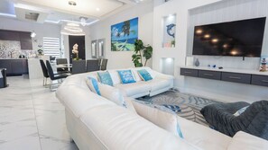 A modern living room with a sectional sofa, a flat-screen TV, and a dining area with chairs and a table. - Sleeps 12, Pool, Walk to Beach (South Padre Island)