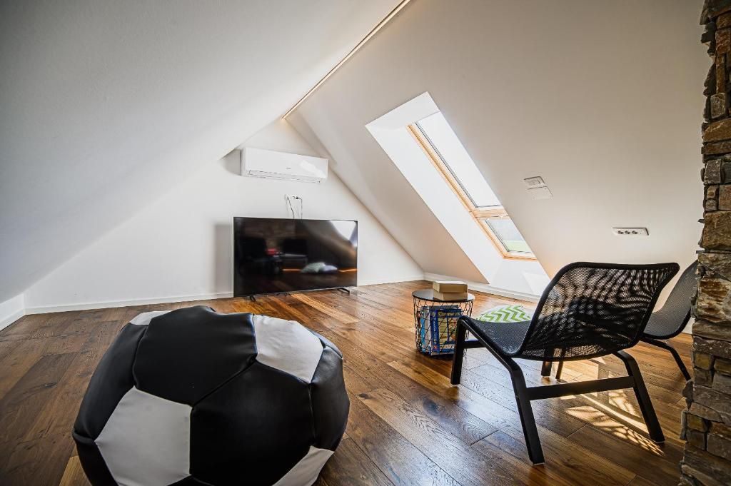 A modern loft bedroom with a sloped ceiling, a skylight, a flat-screen TV, a black and white bean bag, and a black mesh chair.