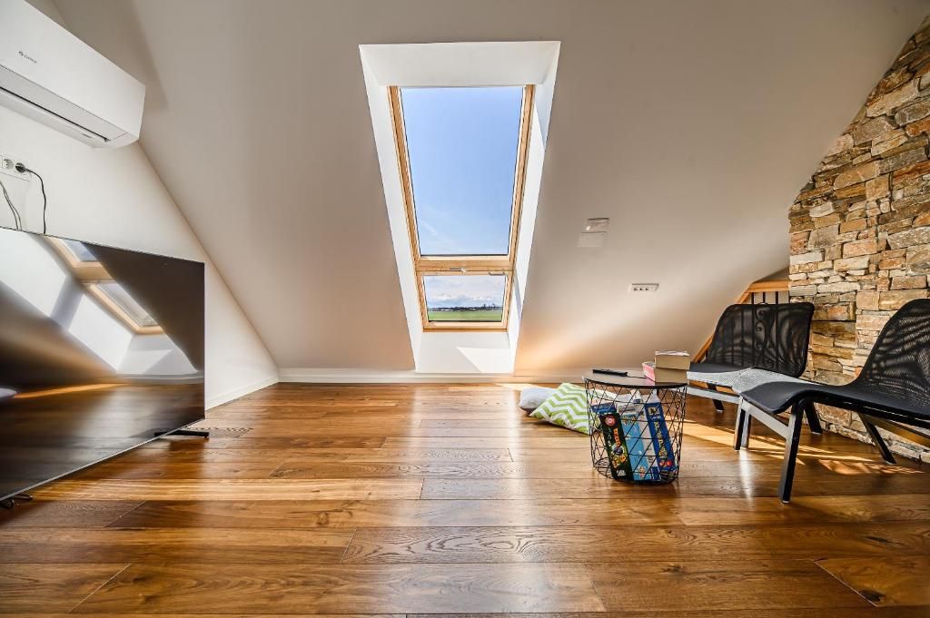 Une pièce avec un plafond en pente, un puits de lumière, un plancher en bois, une chaise noire, un mur blanc orné d’un motif de pierres et une petite table sur laquelle se trouve un objet coloré.