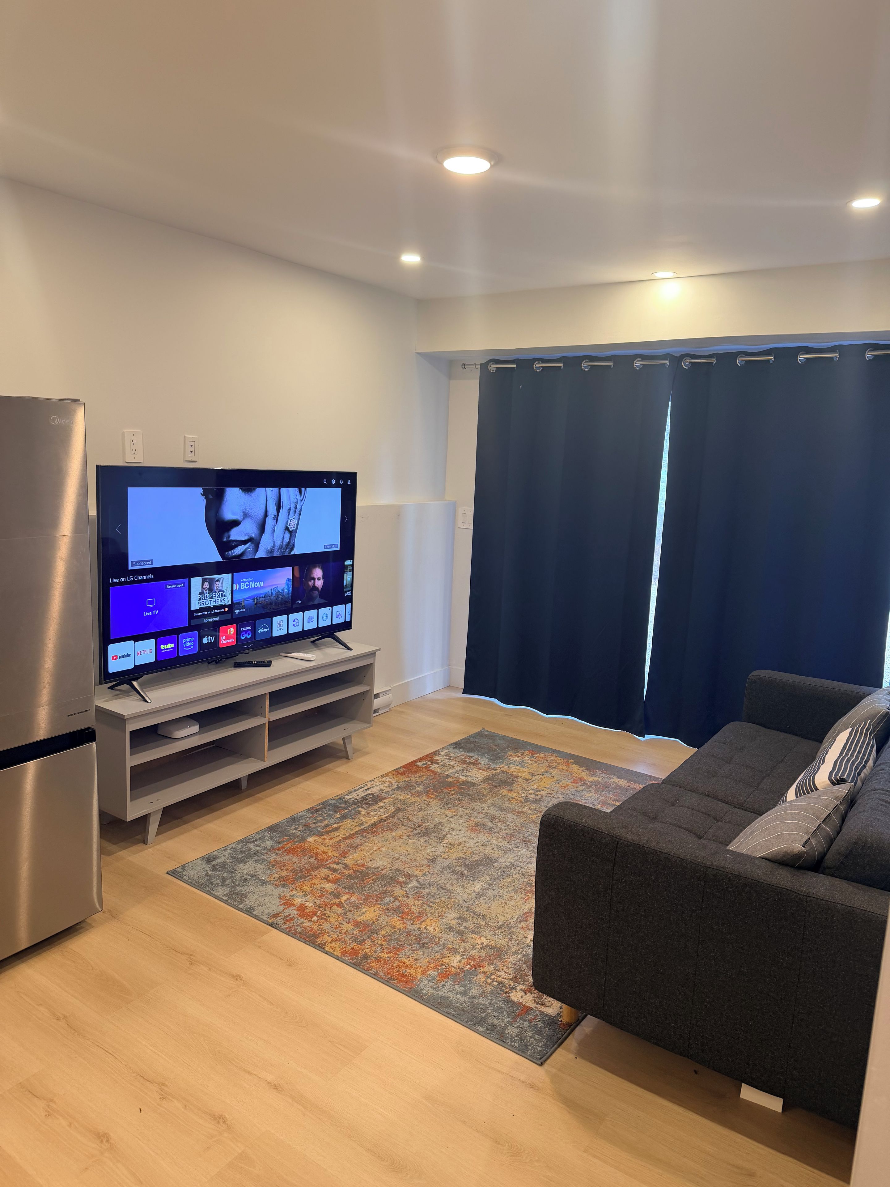 A modern living room with a flat-screen TV, a sofa, a rug, and a refrigerator.