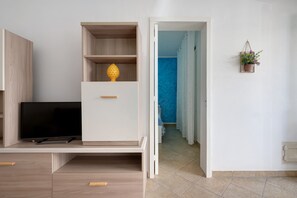 Une pièce avec un téléviseur posé sur un meuble, une armoire blanche ornée d’un objet décoratif, et une porte donnant sur un couloir avec un mur bleu.