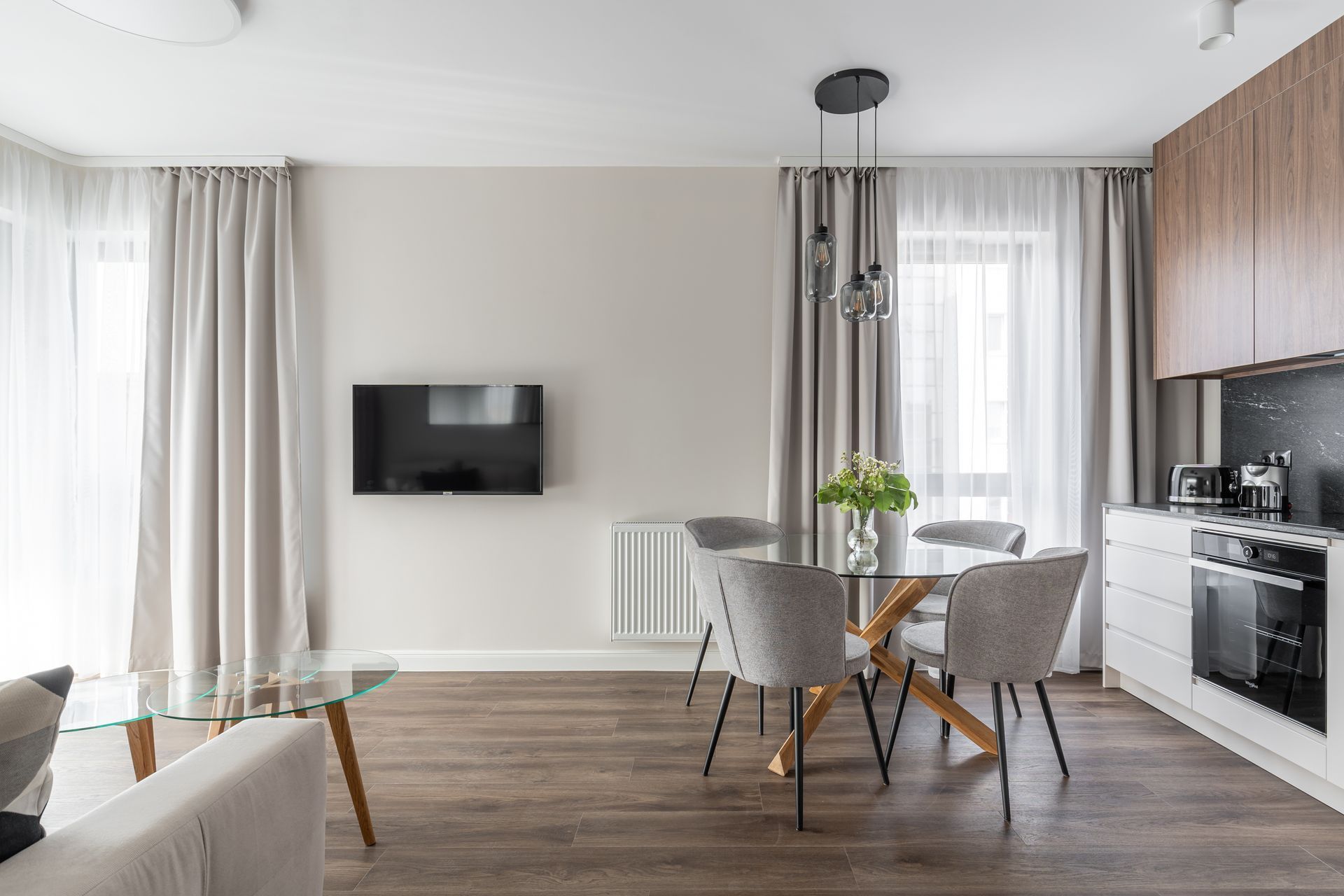 Een moderne woonkamer met een glazen salontafel, grijze stoelen, een flatscreen televisie en een keuken met houten keukenkastjes en een fornuis.