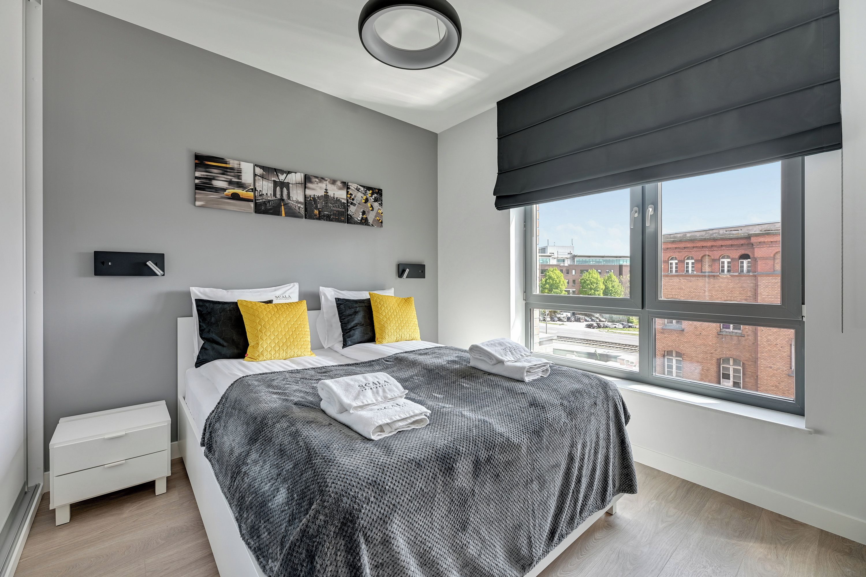 Een moderne slaapkamer met een groot bed, een nachtkastje, een raam met uitzicht op gebouwen en een stadsgezicht schilderij aan de muur.