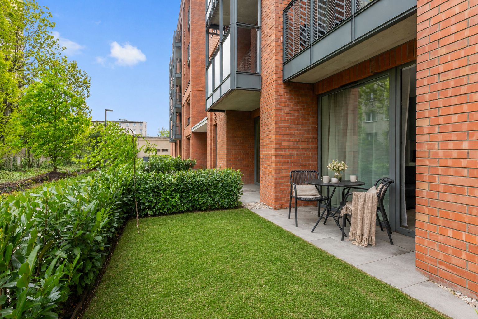 Een modern gebouw met een bakstenen gevel, een kleine tuin met gazon en hagen, en een terras met tuinmeubilair.