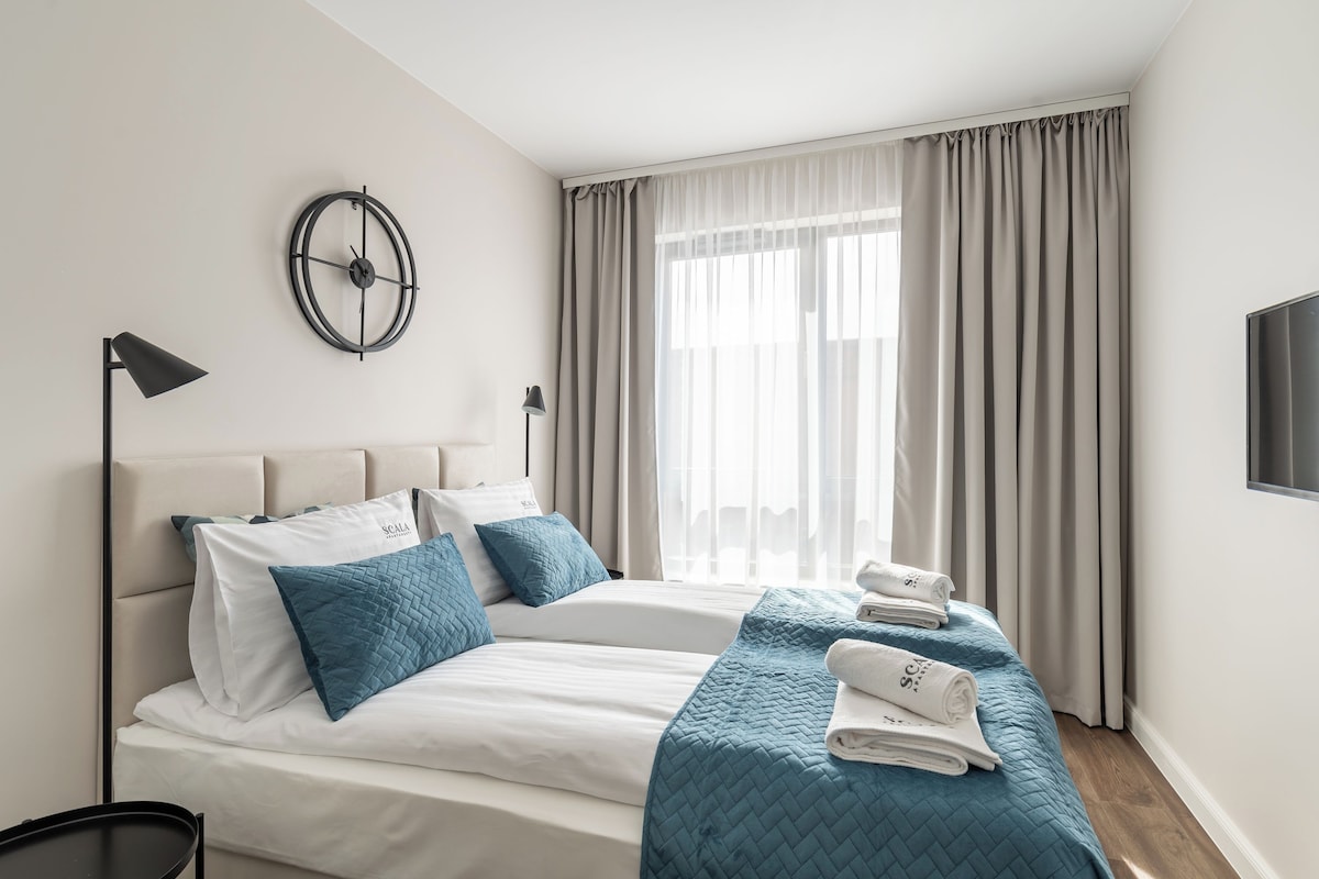 Una habitación de hotel moderna con dos camas, un reloj de pared redondo, un televisor y un gran ventanal con cortinas.