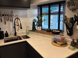 A kitchen with a sink, a window with blinds, a potted plant, and various kitchen utensils.