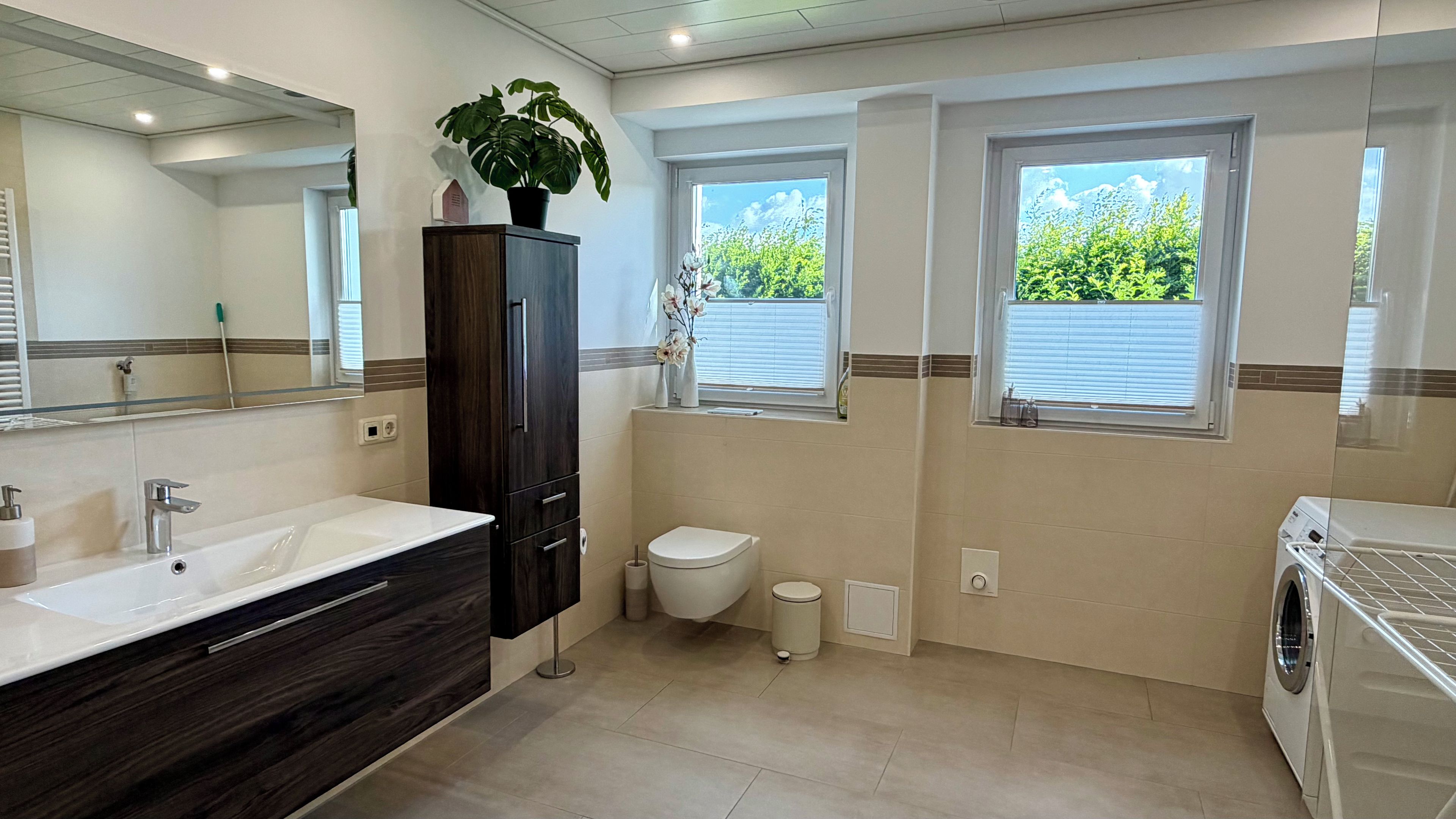 A modern bathroom with a large mirror, dark wood cabinetry, a white sink, a toilet, and a washing machine.