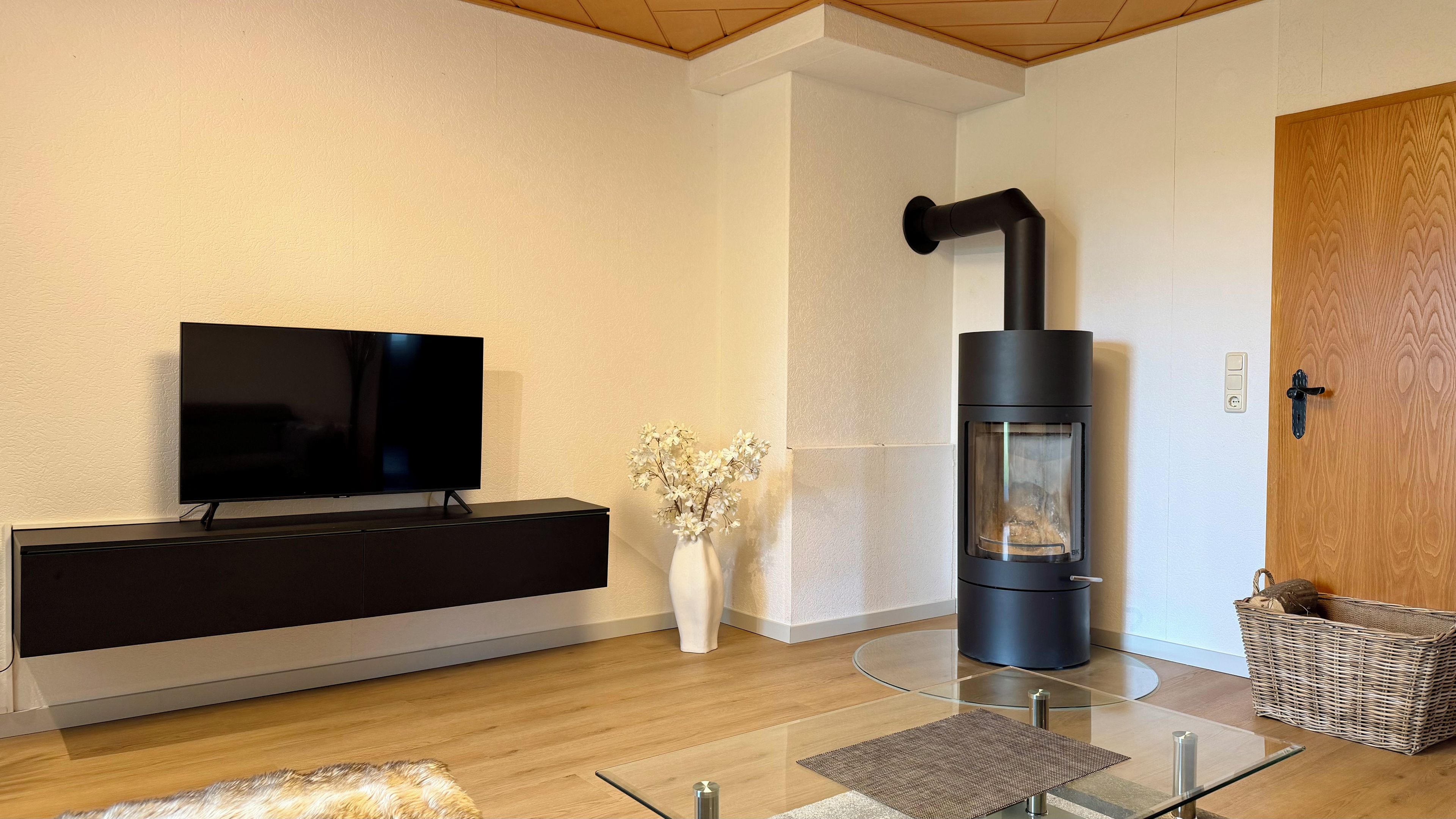 Un salon moderne avec un téléviseur à écran plat, une cheminée noire, un vase blanc orné de fleurs et un panier en osier.